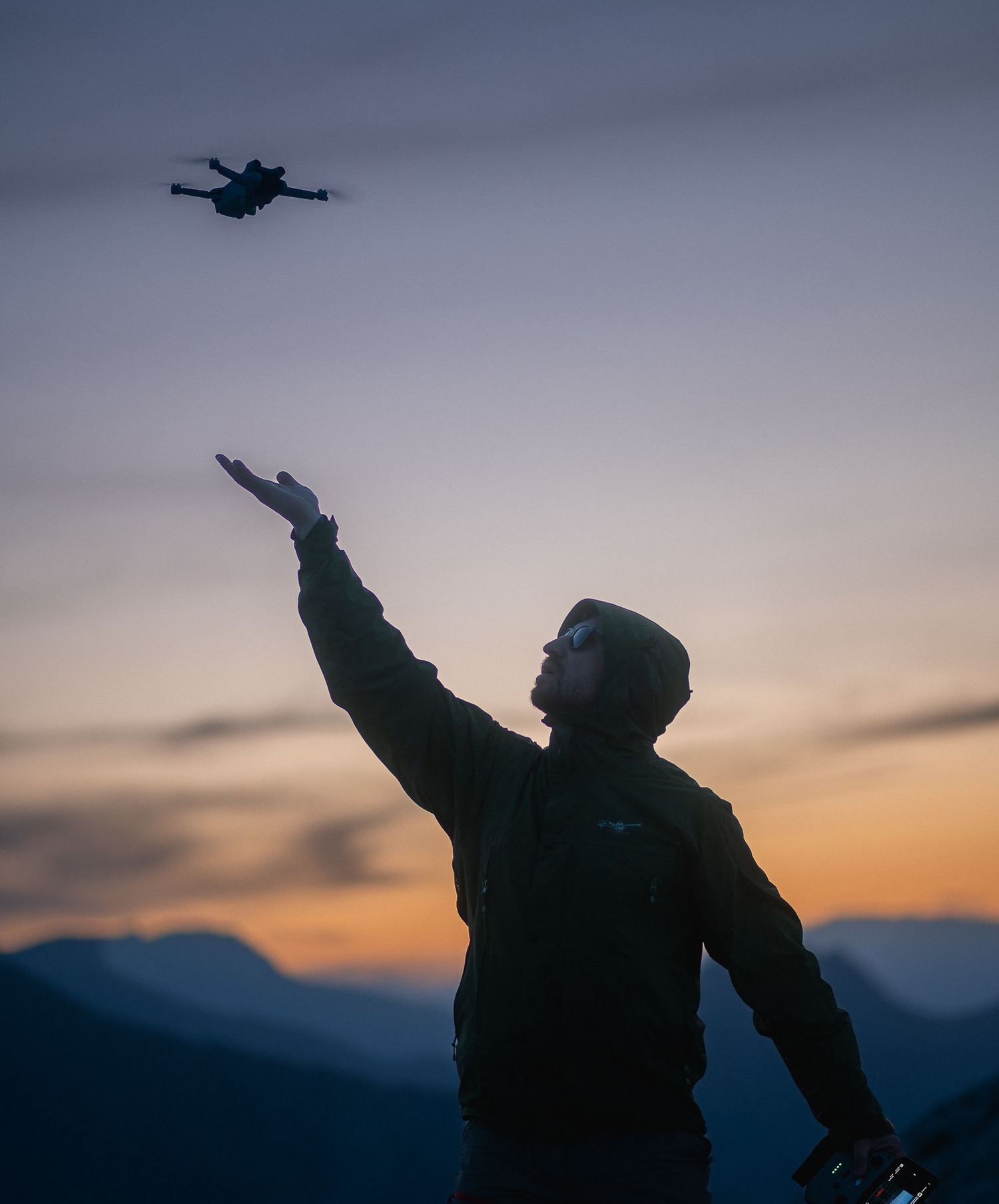 A drone pilot in sunglasses reaches up toward his drone against a dusk sky, controller in hand.