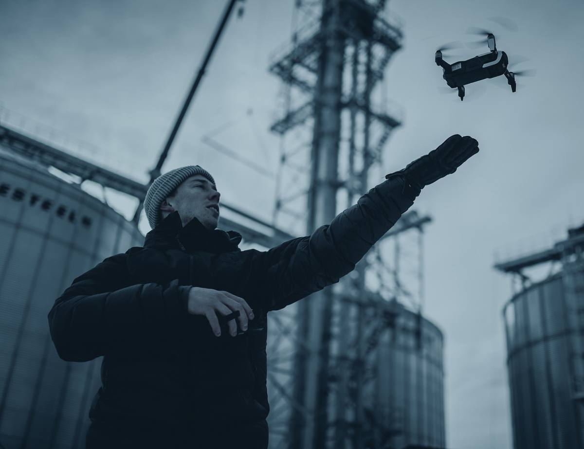 A pilot in a winter jacket launches a small drone from his hand beside grain silos.